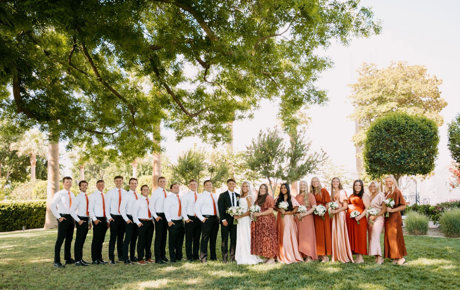 A large wedding party in coordinated attire poses outdoors under green trees.