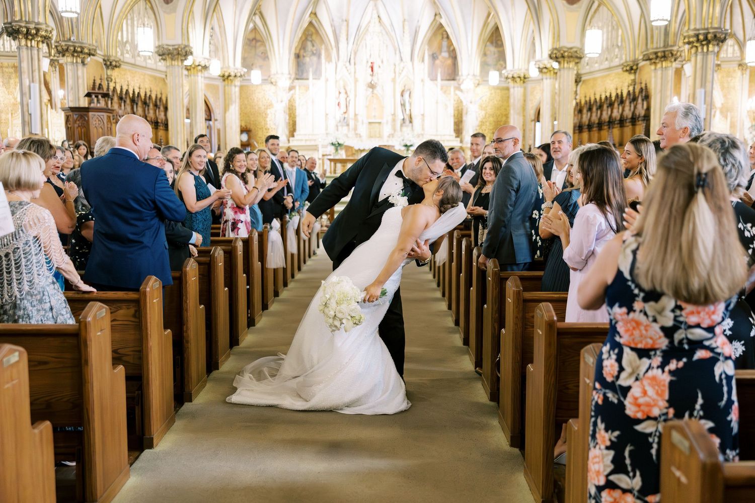 A couple shares a romantic dip kiss in the center aisle of an ornate cathedral church surrounded by guests.