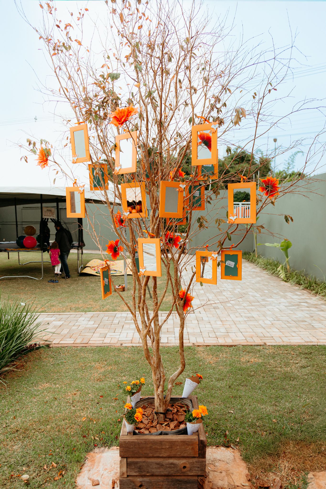 Outdoor garden decor setup with hanging orange lanterns and wooden planter boxes along a paved walkway against a grey wall.