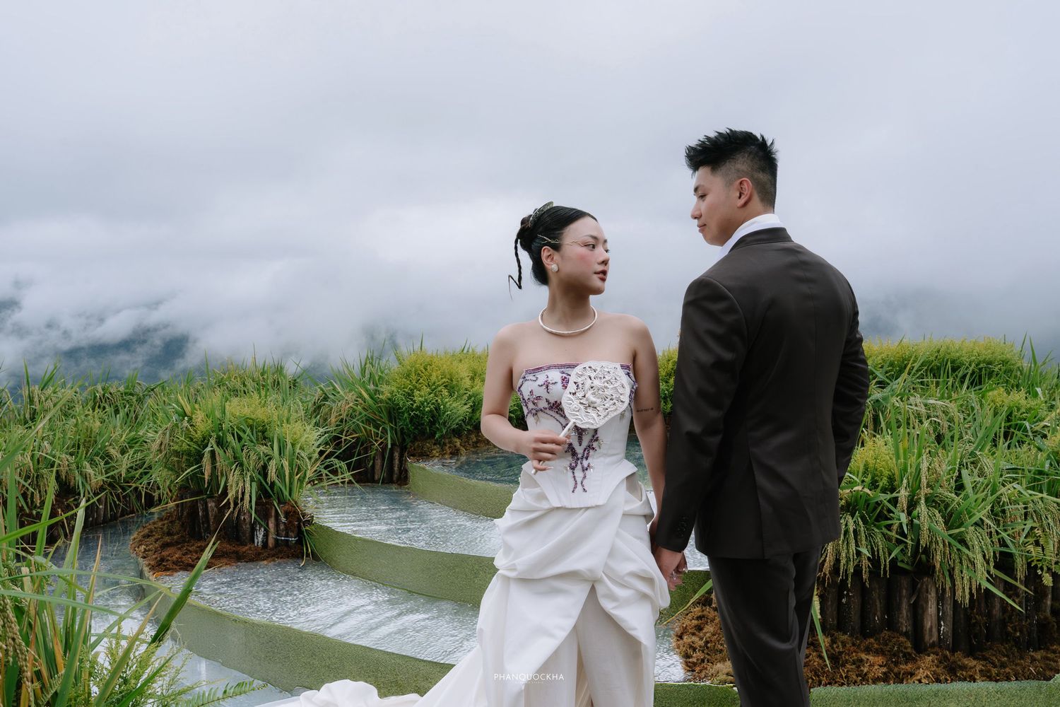 Couple in wedding attire holding hands among lush green rice terraces with misty mountain backdrop.