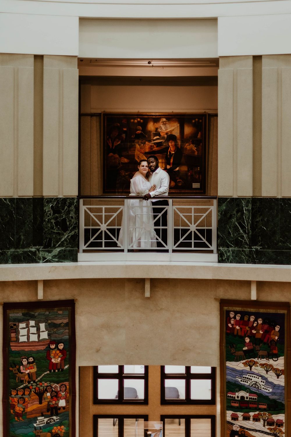 Two people stand intimately together on a balcony overlooking decorative posters in an indoor mall or theater setting.