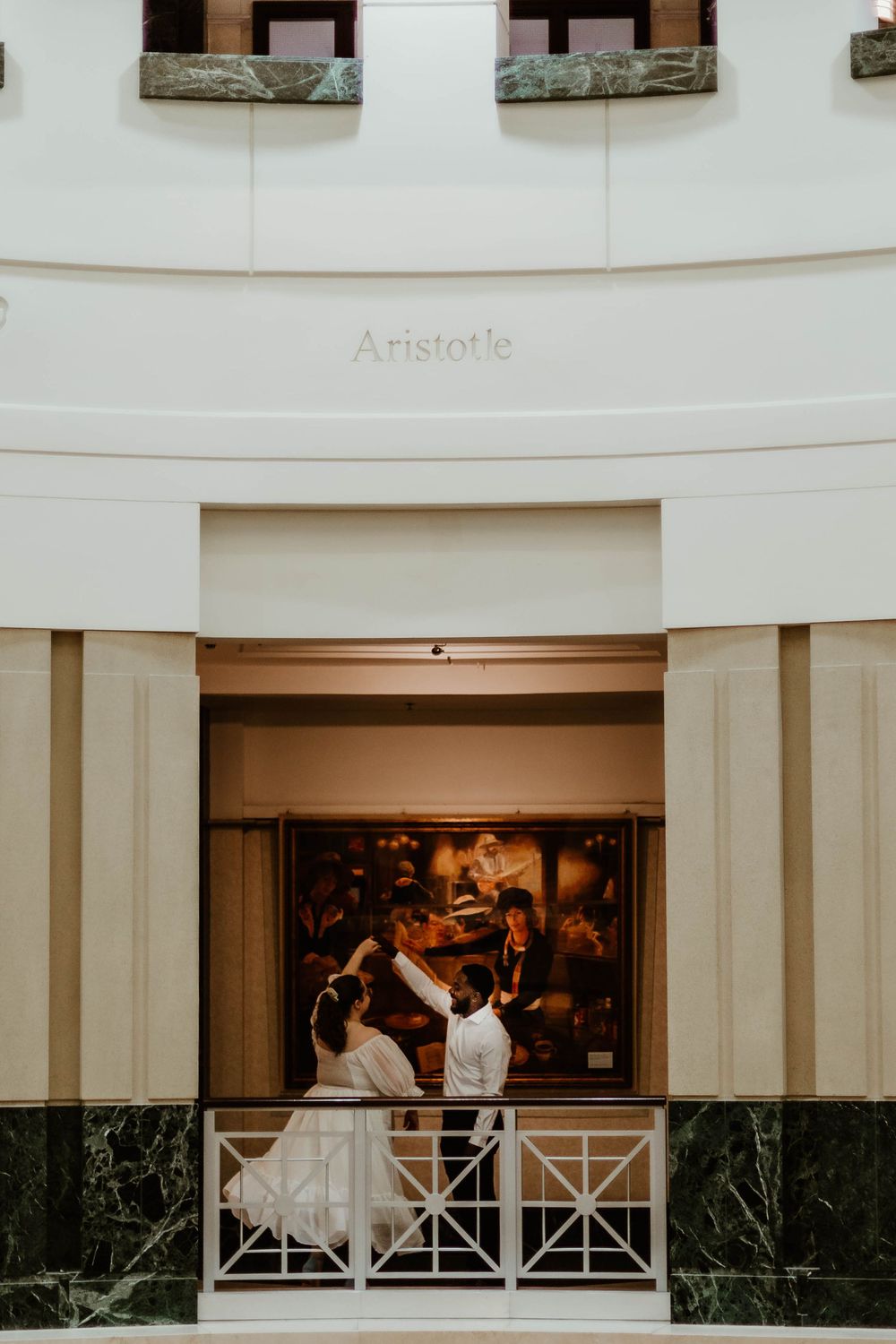 A couple shares an intimate moment on a balcony overlooking an elegant indoor venue.