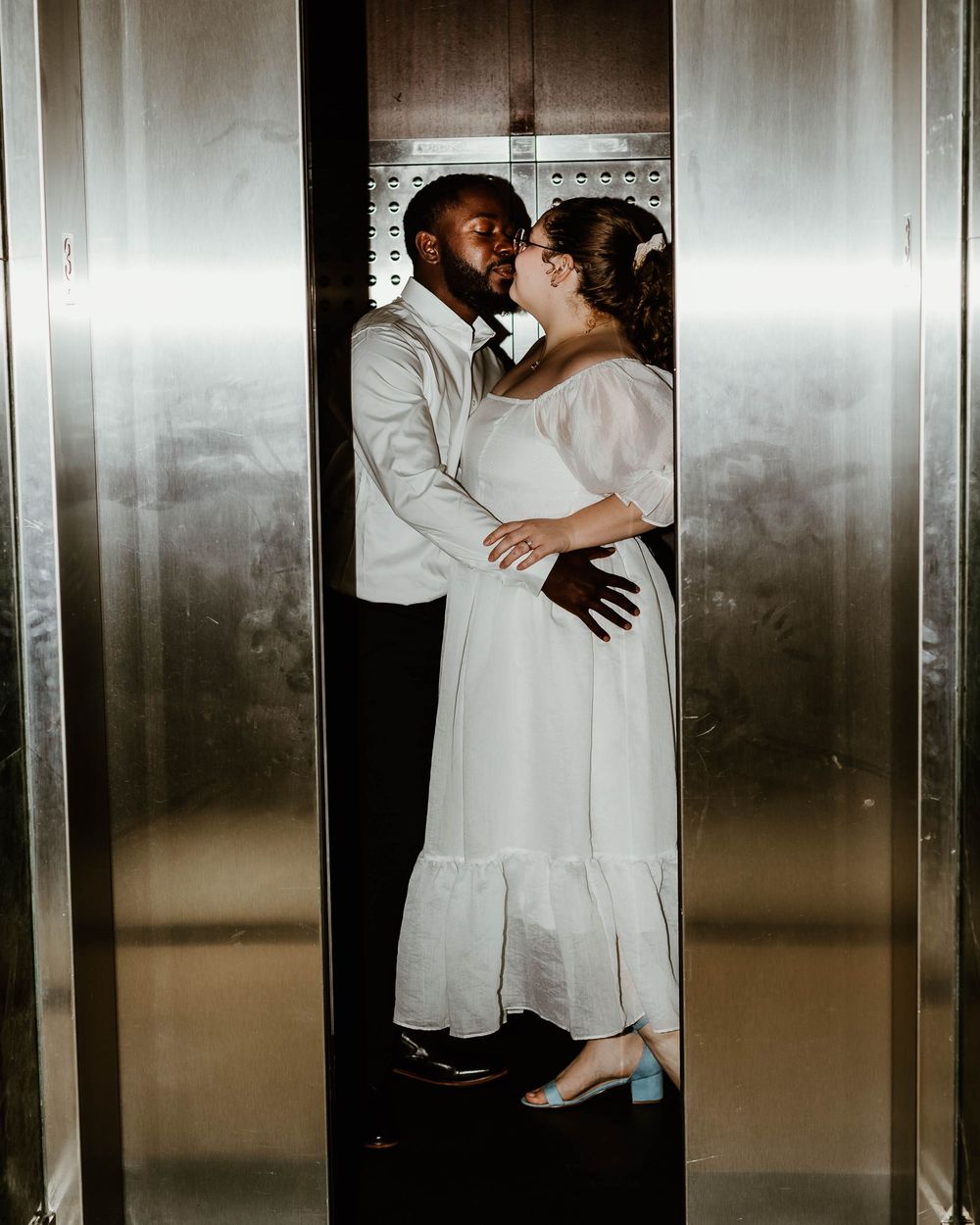 A couple embraces intimately in a series of elevator photos wearing white clothing against a metallic background.