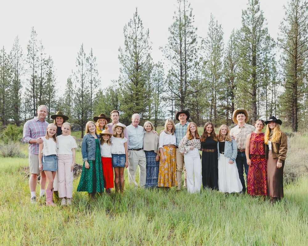 Large family group portrait outdoors in a green meadow with tall pine trees, dressed in casual western-style attire.