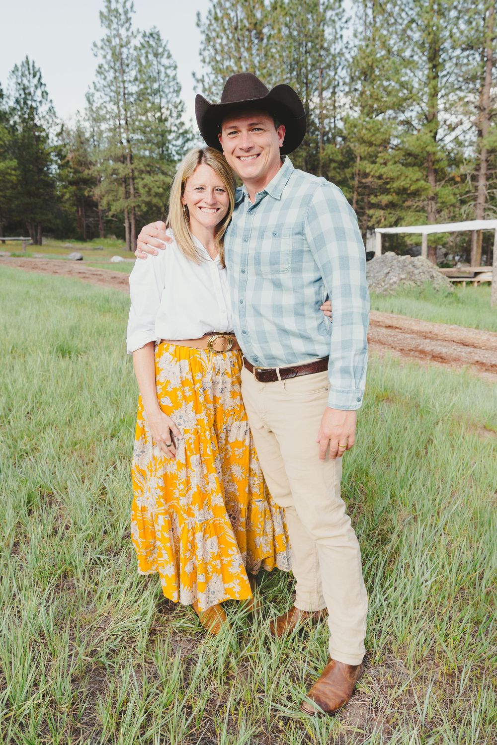 A smiling couple poses outdoors on a green field, woman in yellow floral skirt, man in plaid shirt and cowboy hat.