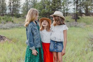 Three young girls in cowboy hats laughing together in a green meadow, wearing colorful summer outfits.