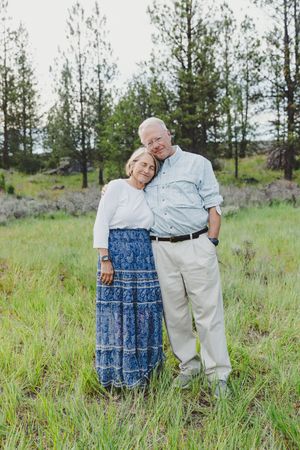 Elderly couple embracing in a green meadow with pine trees, woman in blue skirt leaning on man in light blue shirt.
