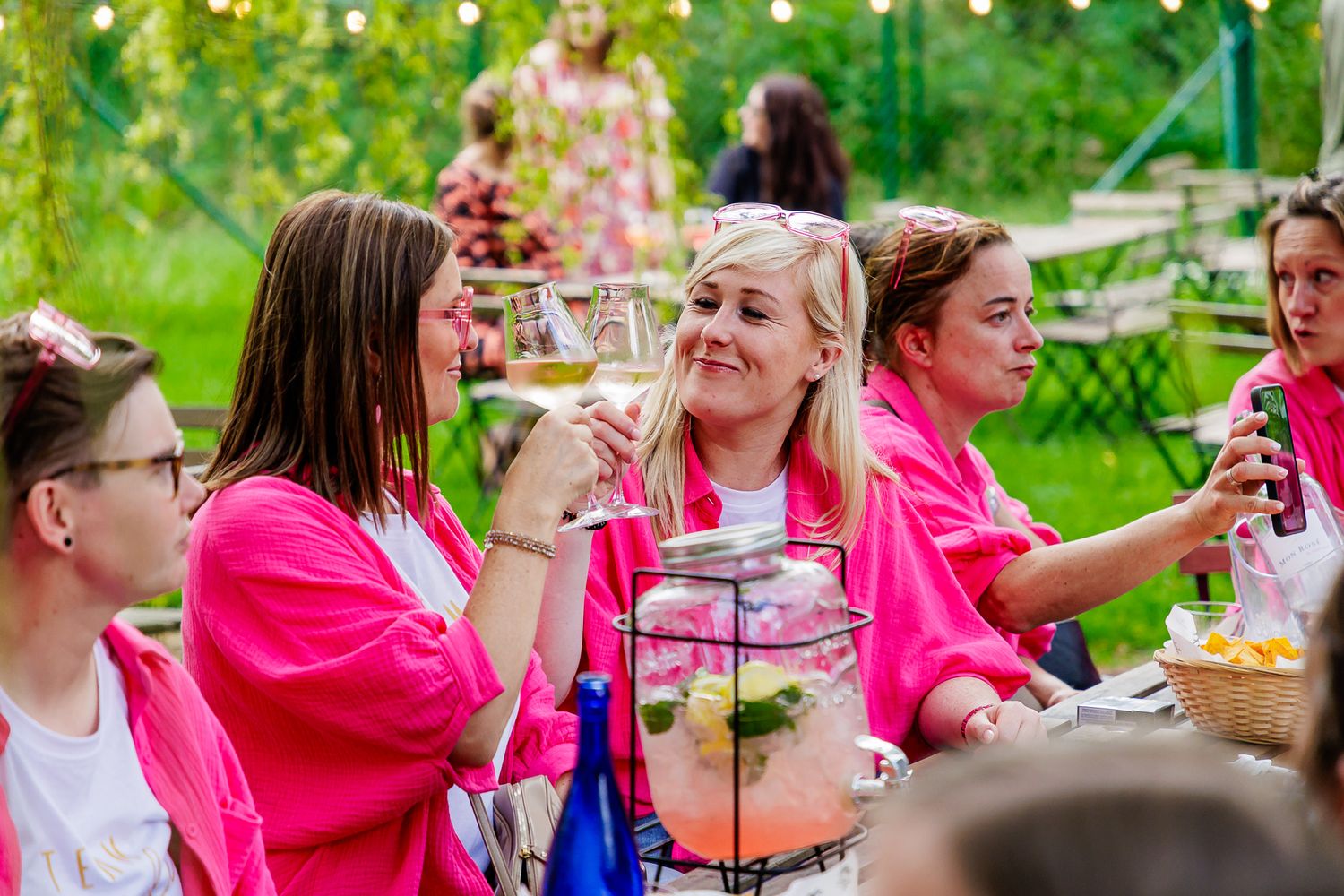 A large group in bright pink shirts socializes at an outdoor garden party with food and drinks spread across tables.