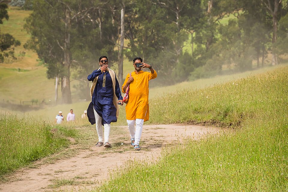 Group of people in casual attire walking together along a scenic dirt path through green grass on a bright sunny day.