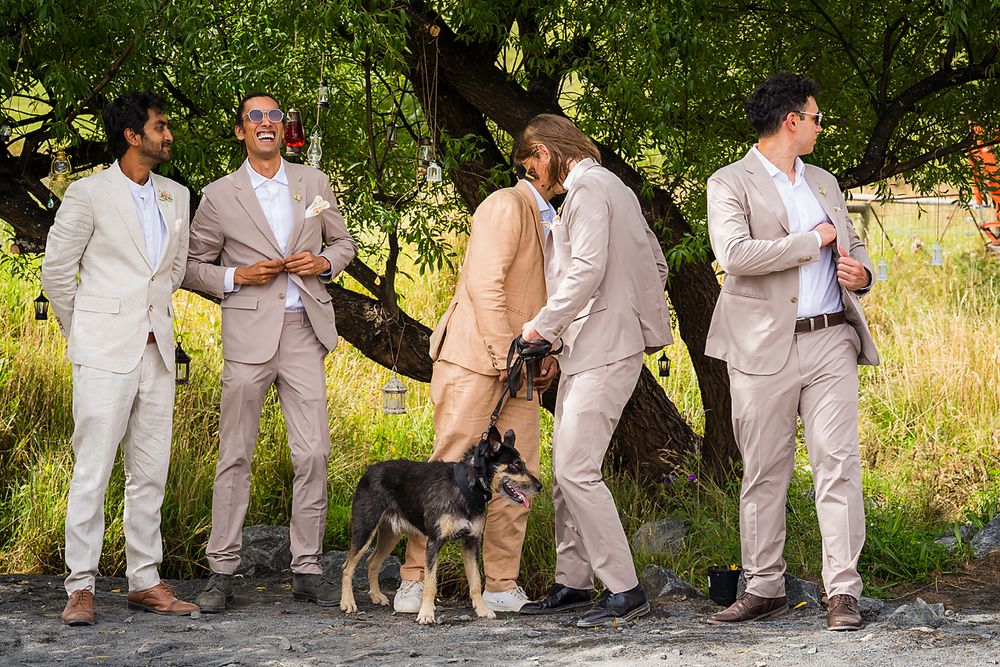 Wedding party in light colored attire walks in a line through lush outdoor garden setting, creating a playful conga line.