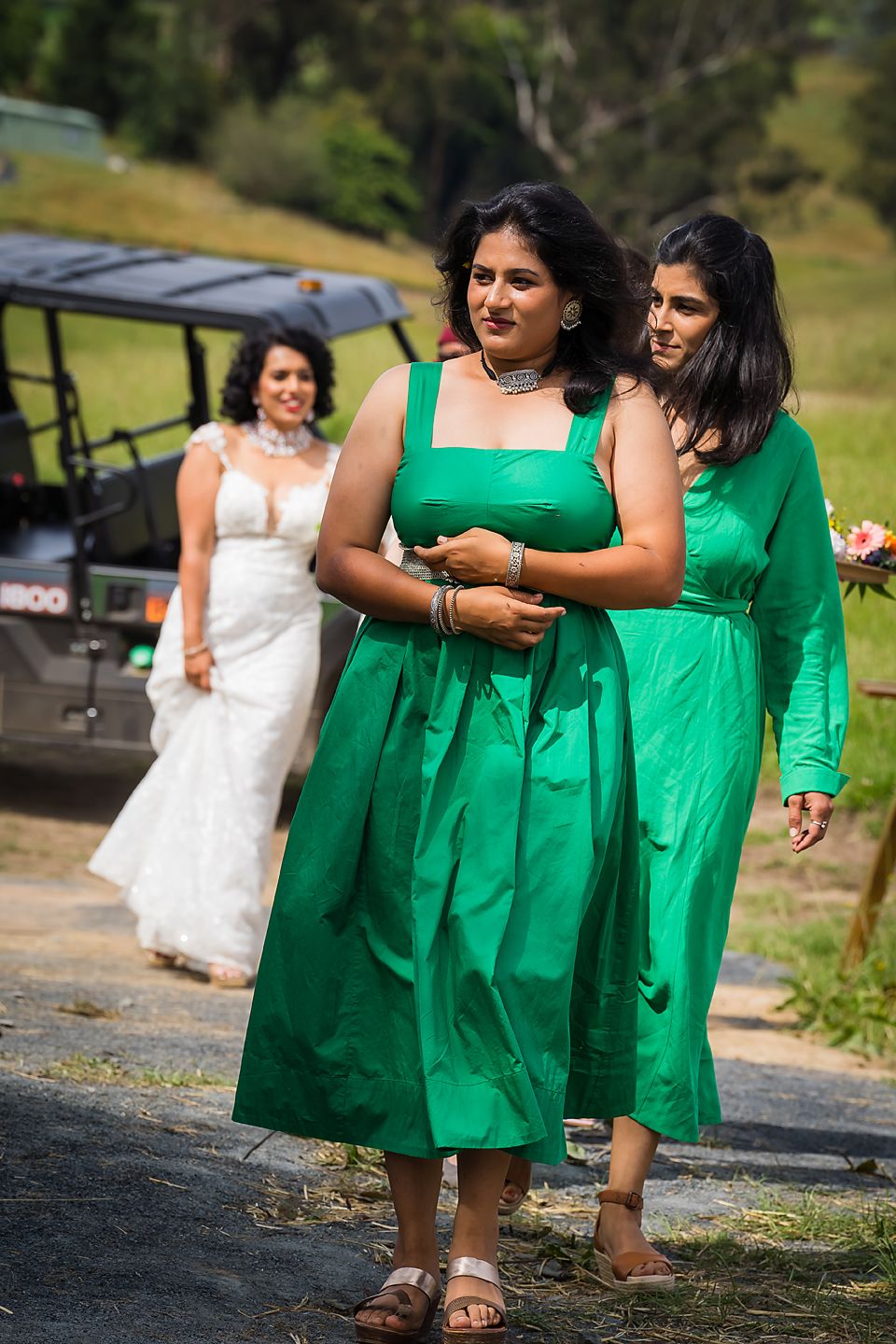 Bridesmaids in vibrant emerald green dresses walk alongside a bride in white during an outdoor wedding celebration.