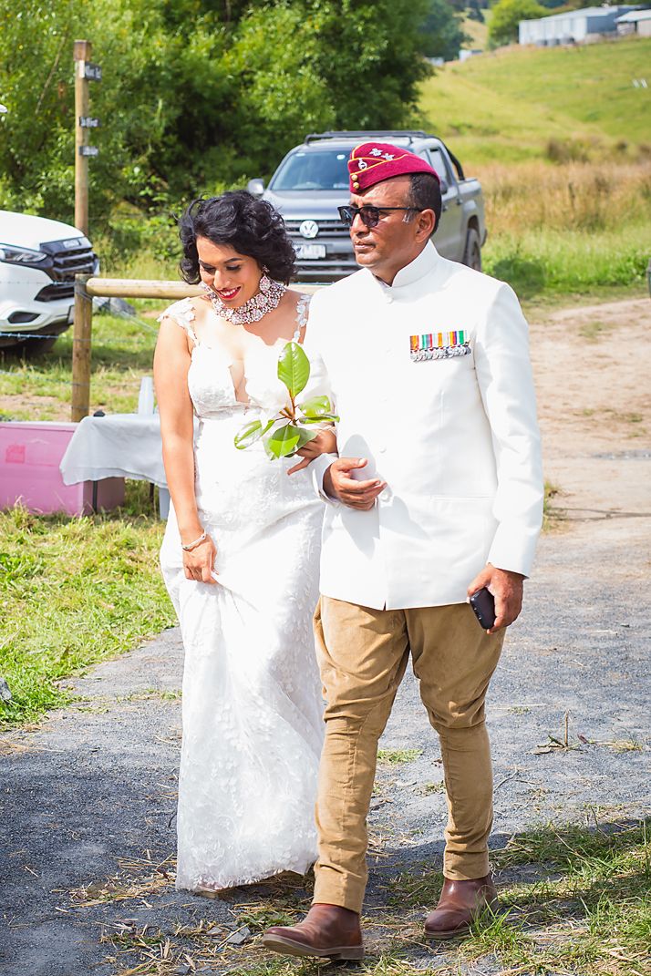 A wedding couple in white and khaki attire stand together on a path surrounded by greenery during a summer celebration.