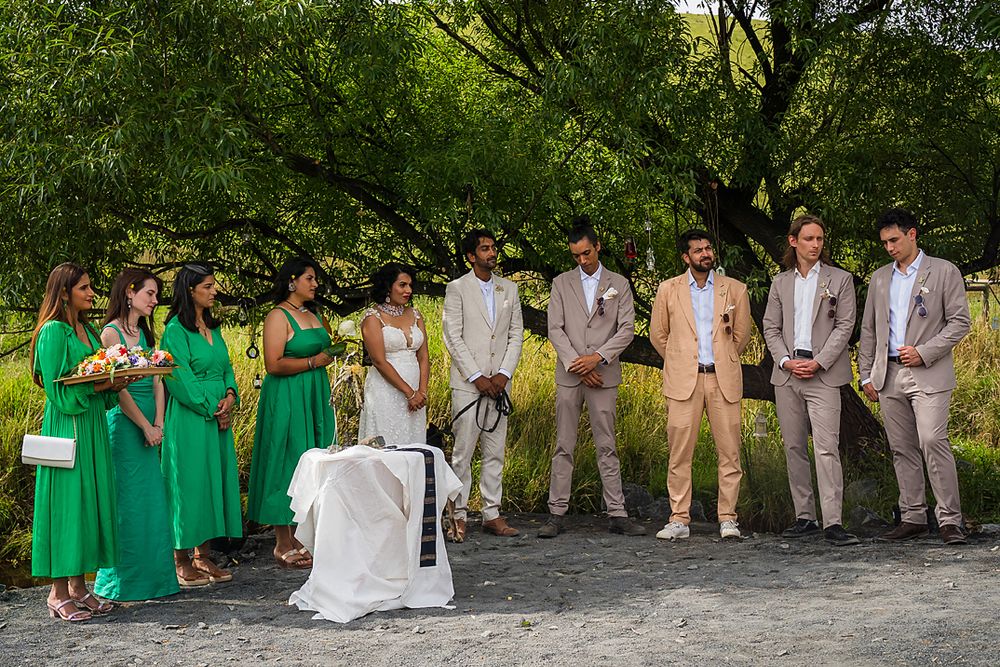 Wedding party members in bright emerald green dresses and tan suits gather outdoors under lush trees.