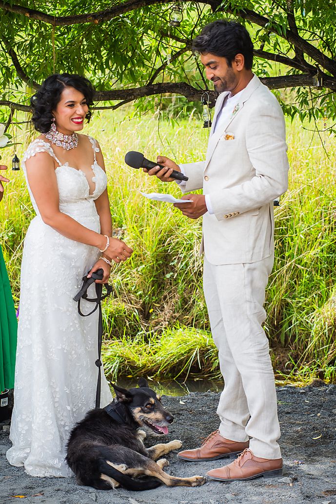 A couple in white wedding attire share a moment outdoors with their black dog resting at their feet.