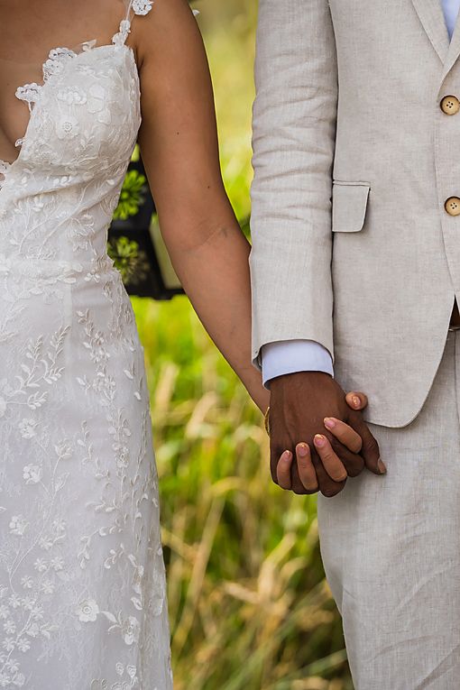 Close-up view of bride in white lace dress and groom in beige suit holding hands during outdoor wedding ceremony.