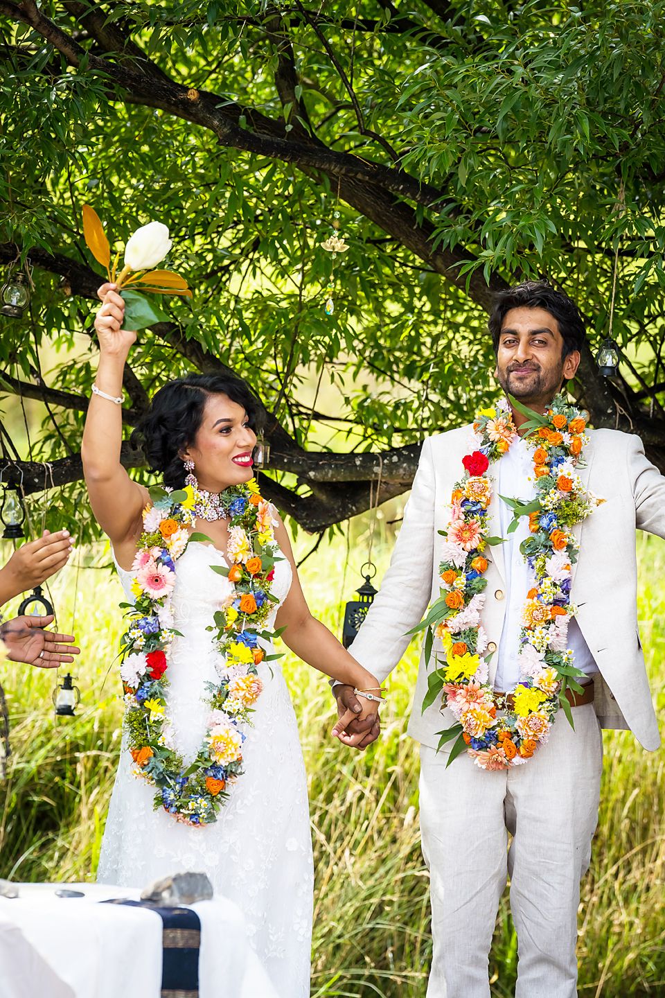 Joyful couple wearing white outfits and floral leis celebrate with dance moves under lush green tree canopy.