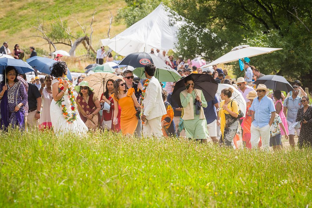 Festival attendees gather under umbrellas and tents at an outdoor summer event with a grassy meadow in the foreground.