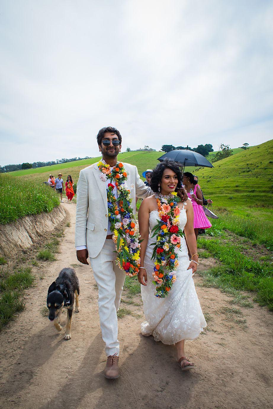 Wedding party walking along a dirt path wearing white attire and colorful leis against green hillside landscape.