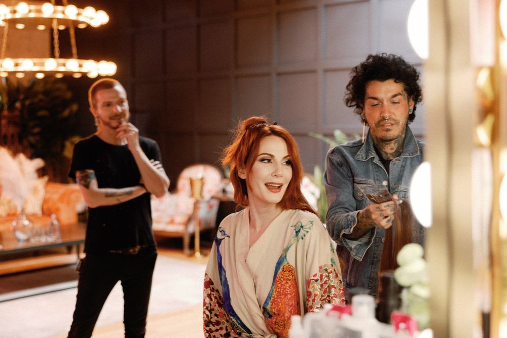 Bride in kimono robe gets hair and makeup done by two stylists in glamorous dressing room.