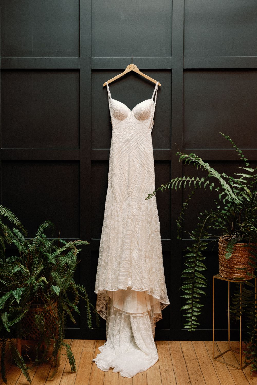 Elegant beaded spaghetti strap wedding gown hanging on wooden hanger against dark paneled wall.