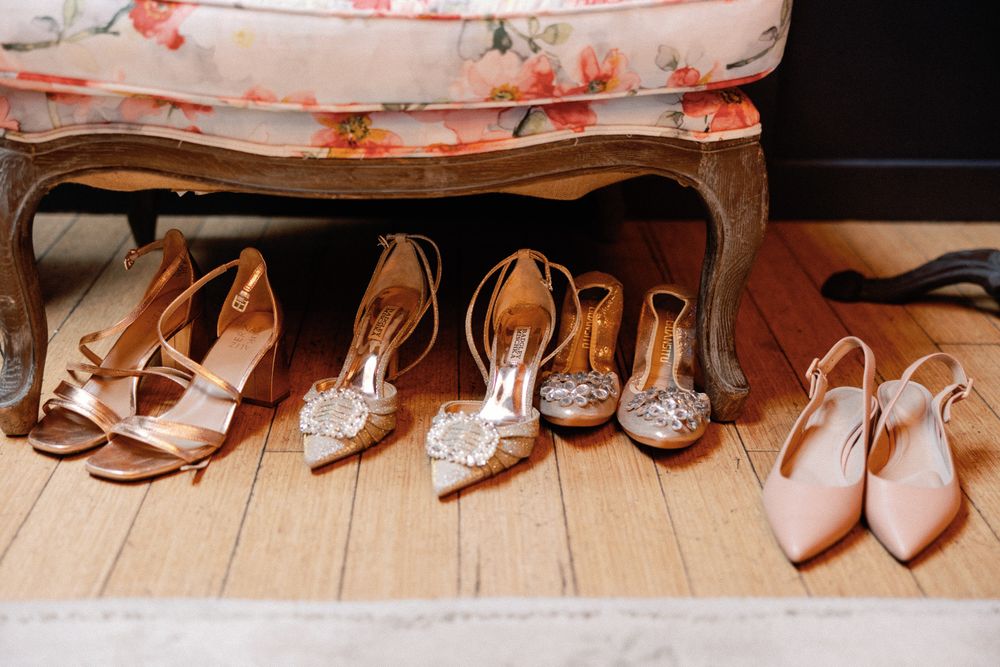 Four pairs of elegant wedding heels arranged on hardwood floor beneath a floral upholstered chair.
