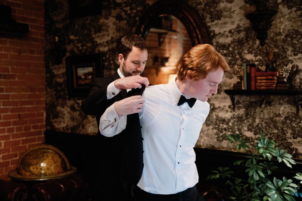 Groom with red hair getting boutonniere pinned by groomsman in dark vintage-styled venue.