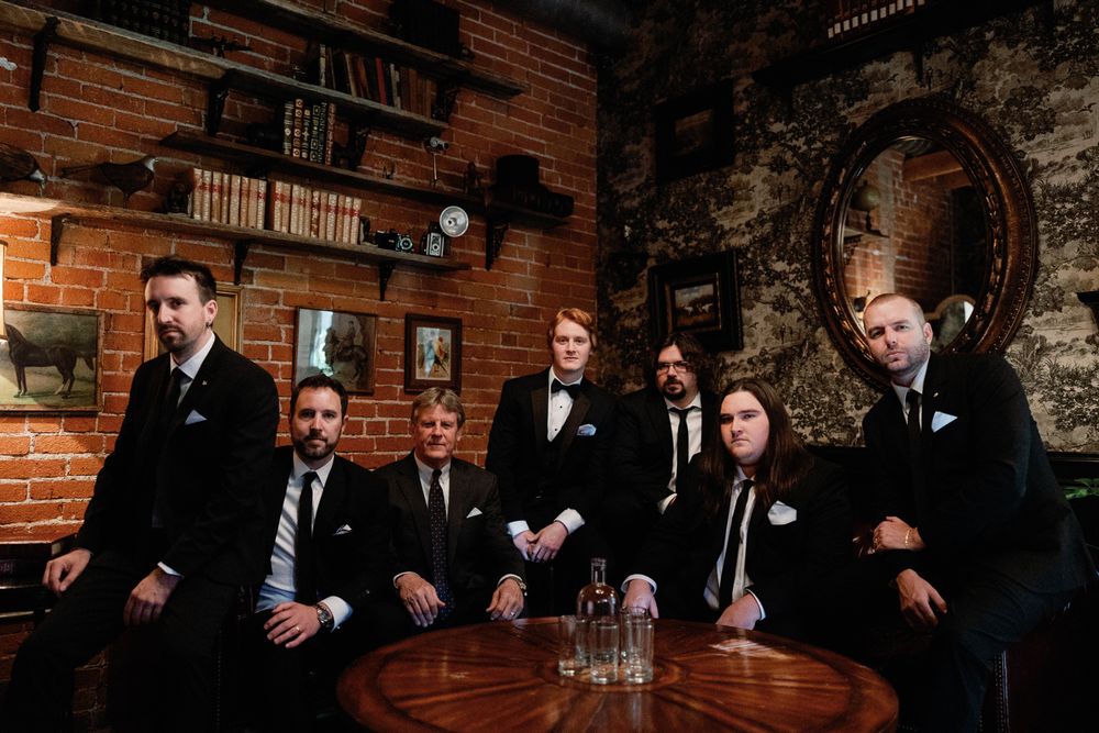 Group of seven groomsmen in black tuxedos posing around wooden table in rustic brick venue.