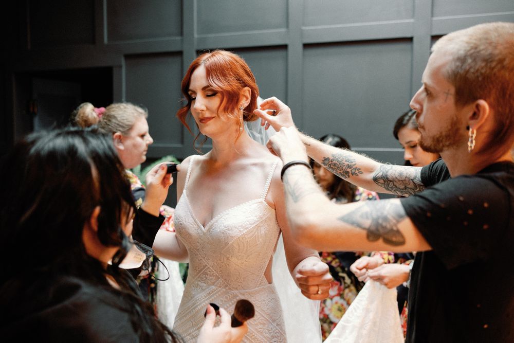 Bride with red hair in white wedding gown being styled by makeup and hair artists before ceremony.