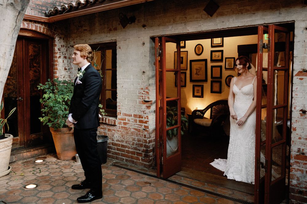 Red-haired groom seeing bride in lace wedding gown for first look in rustic brick courtyard.