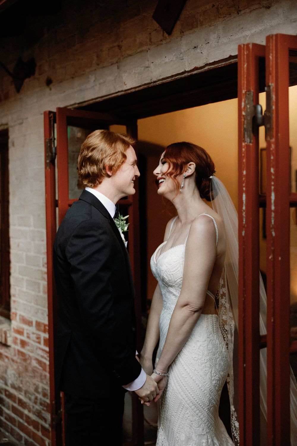 Bride and groom smiling and holding hands in doorway of rustic brick venue on wedding day.