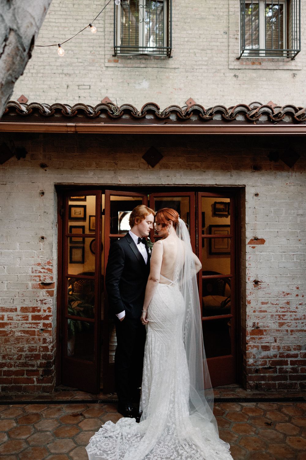 Bride and groom sharing an intimate moment outside a rustic brick building with French doors.
