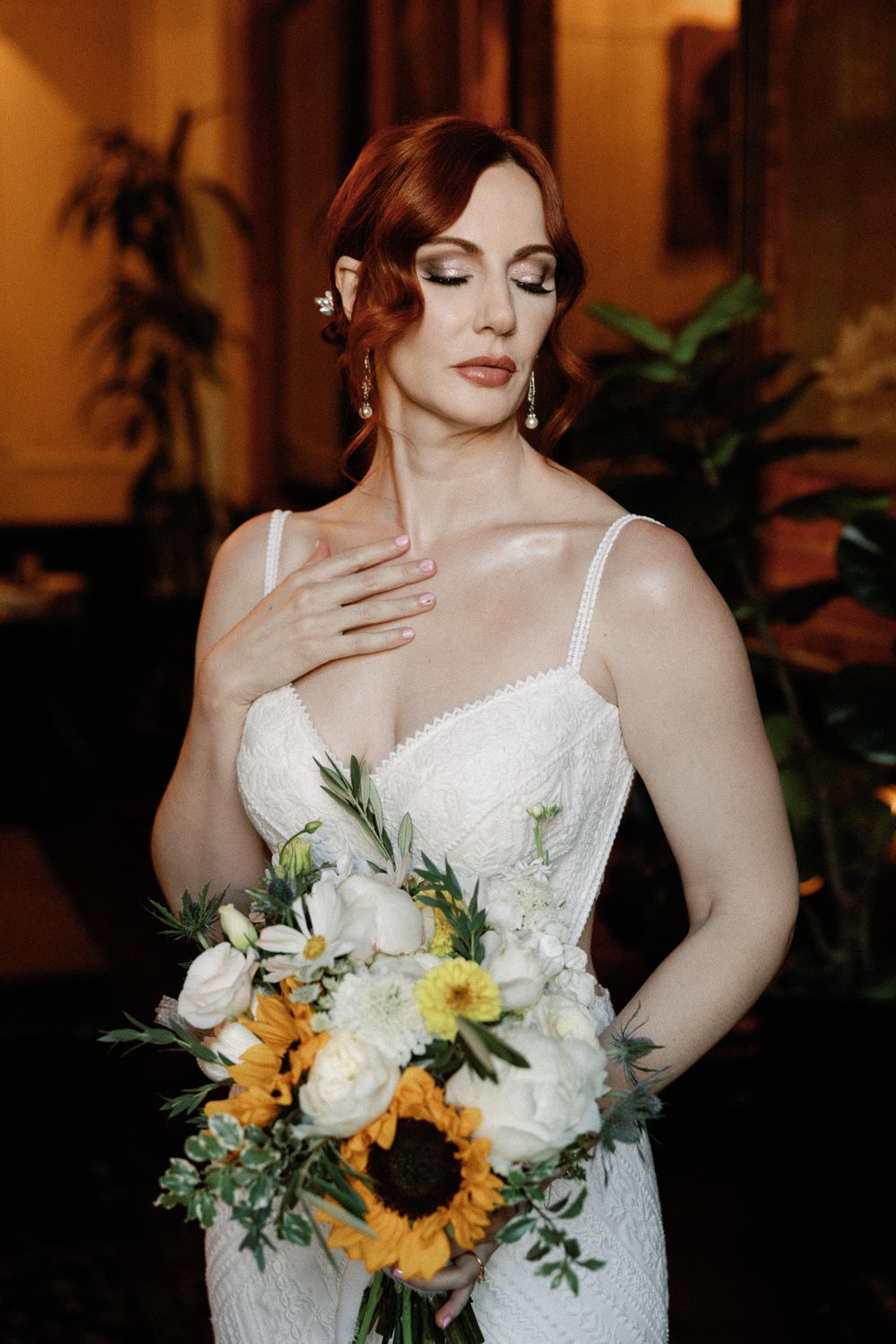 Elegant redhead bride holding a sunflower bouquet in a lace gown with eyes closed.