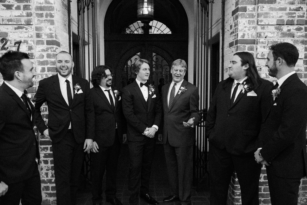 Groom and groomsmen laughing together in black suits outside a brick building, black and white photo.
