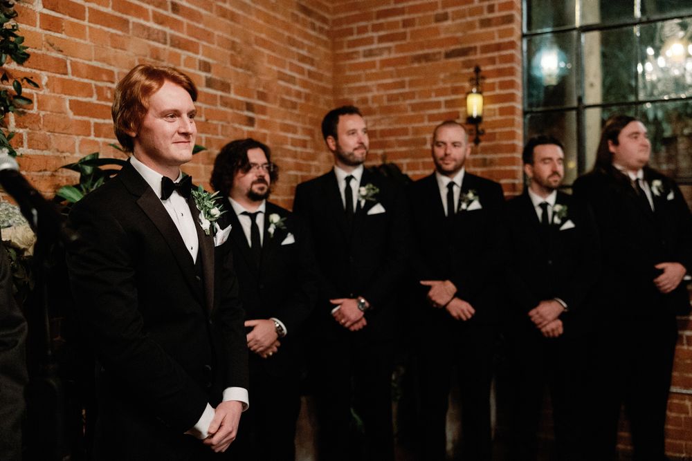 Redhead groom smiling with groomsmen in black tuxedos during wedding ceremony at brick venue.