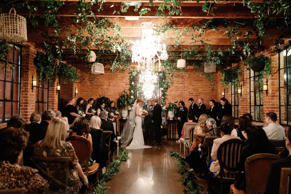 Wedding ceremony in a romantic brick venue adorned with lush greenery garlands and chandeliers.