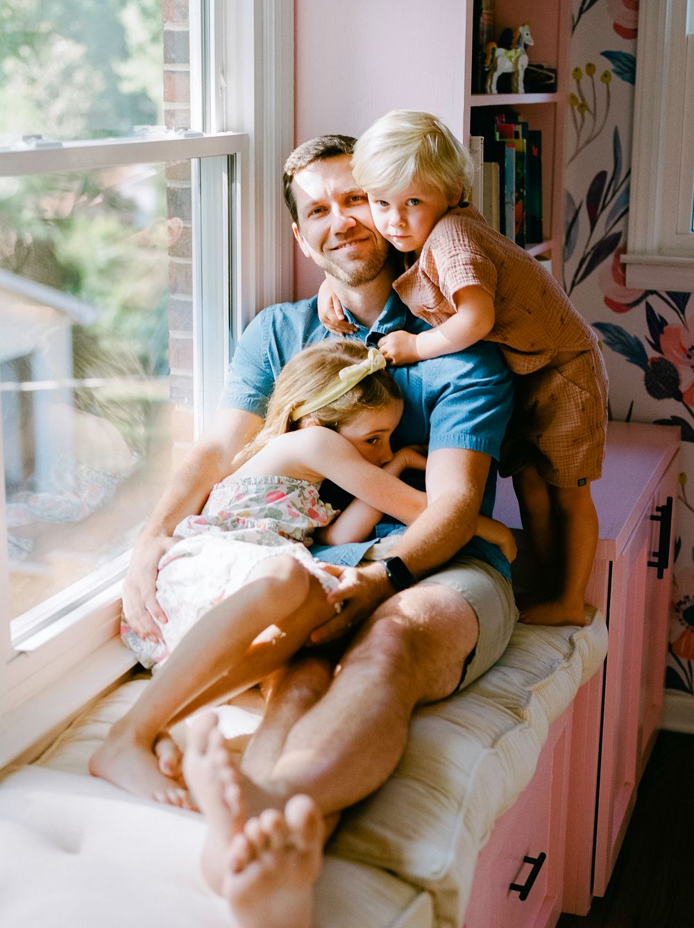Sweet candid moments of family lounging together in pink-walled room by window.