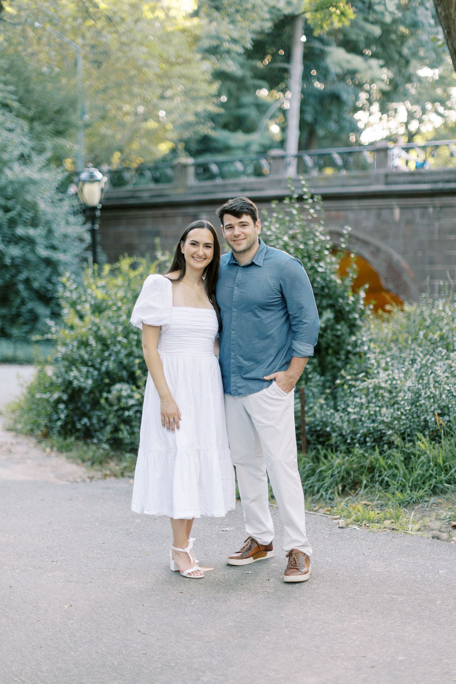 Couple in coordinated white and blue outfits pose together on a garden path during an outdoor photoshoot.