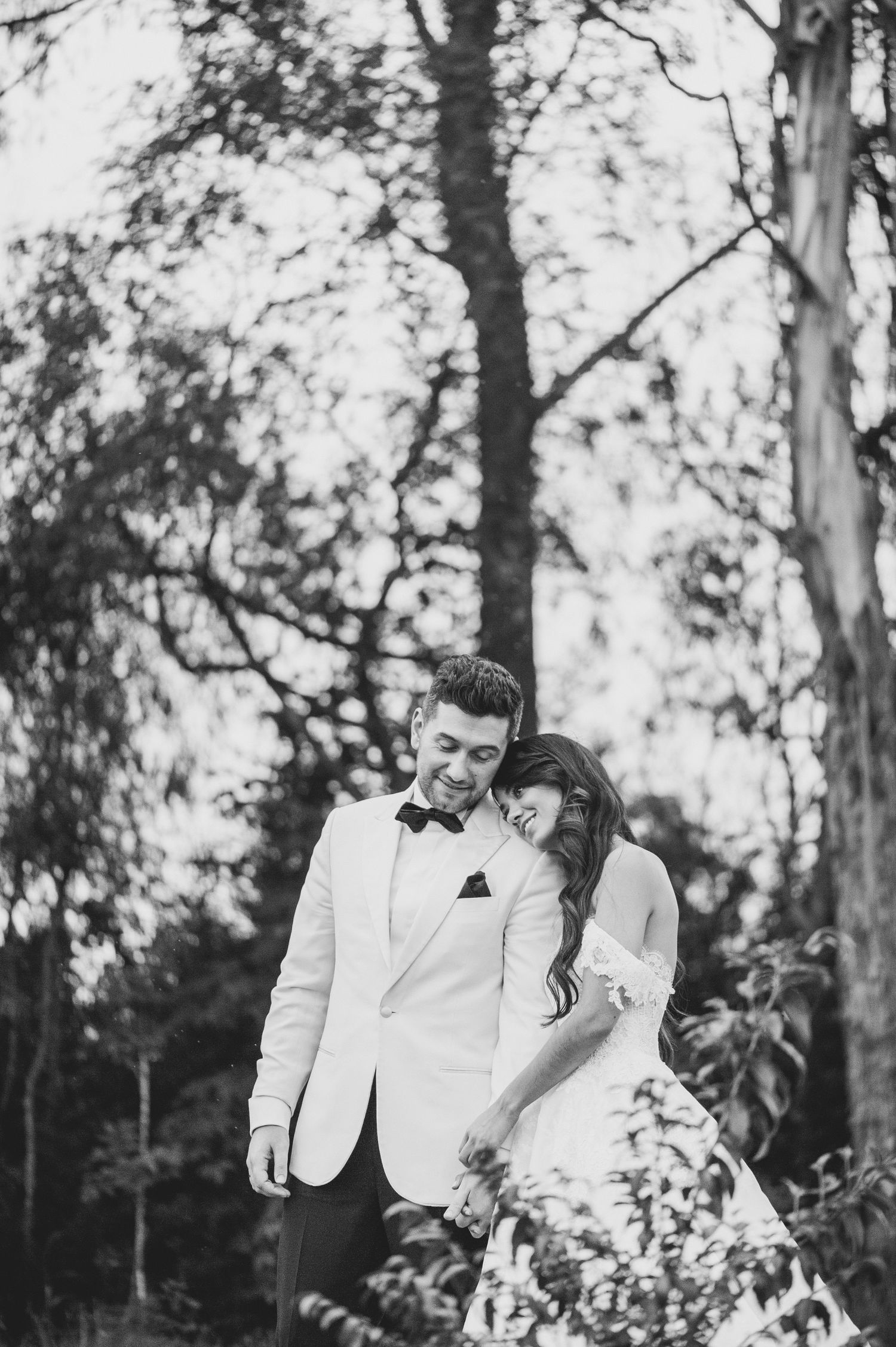 A romantic black and white wedding photo showing a couple embracing outdoors among tall trees in natural lighting.