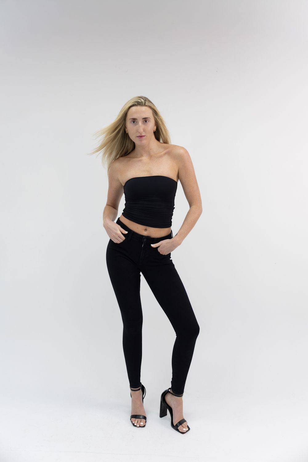 Blonde woman in black tube top and skinny jeans with heels posing on white background.
