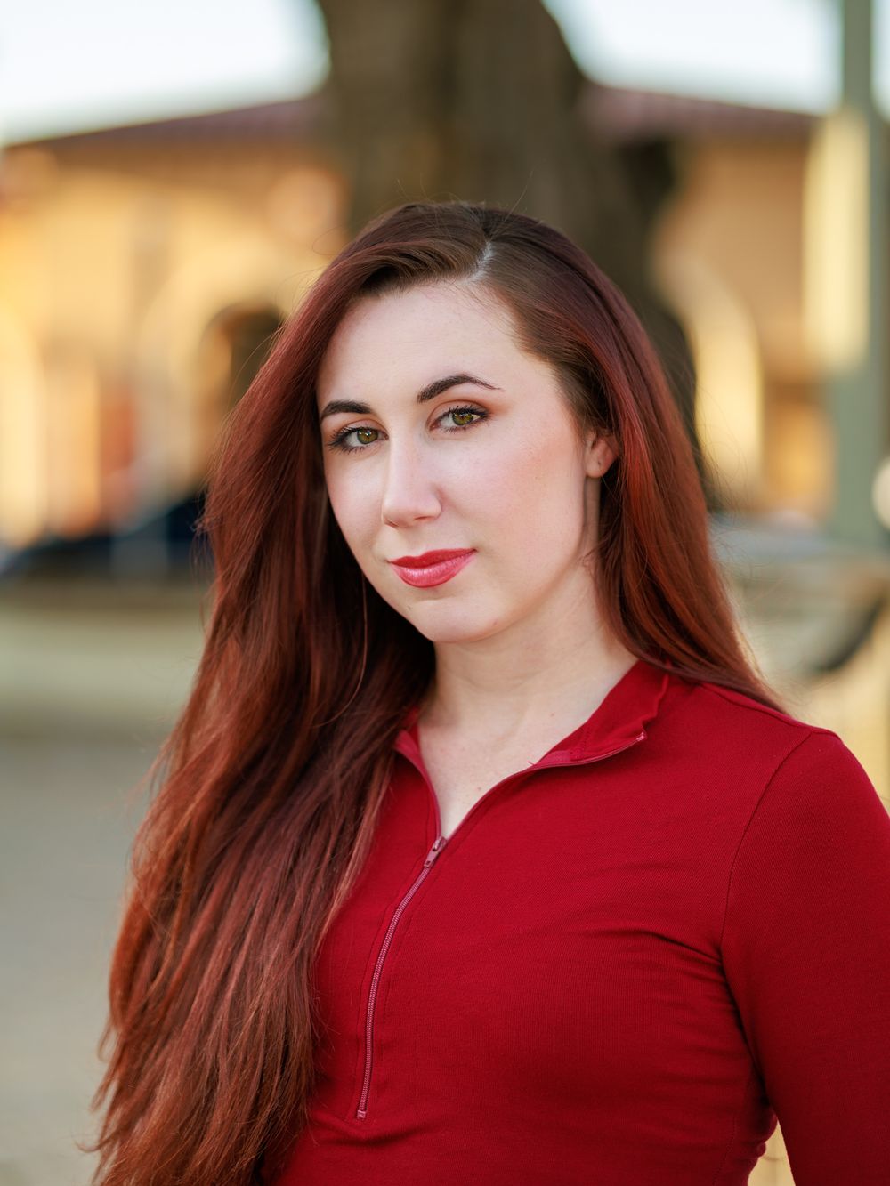 Woman with long auburn hair wearing a red collared top poses outdoors in natural lighting.