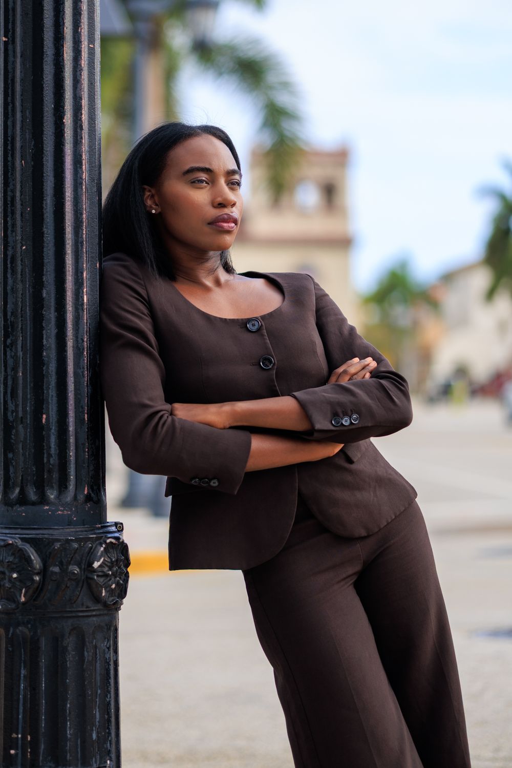 Professional Black woman in brown suit leaning against lamppost outdoors with arms crossed.