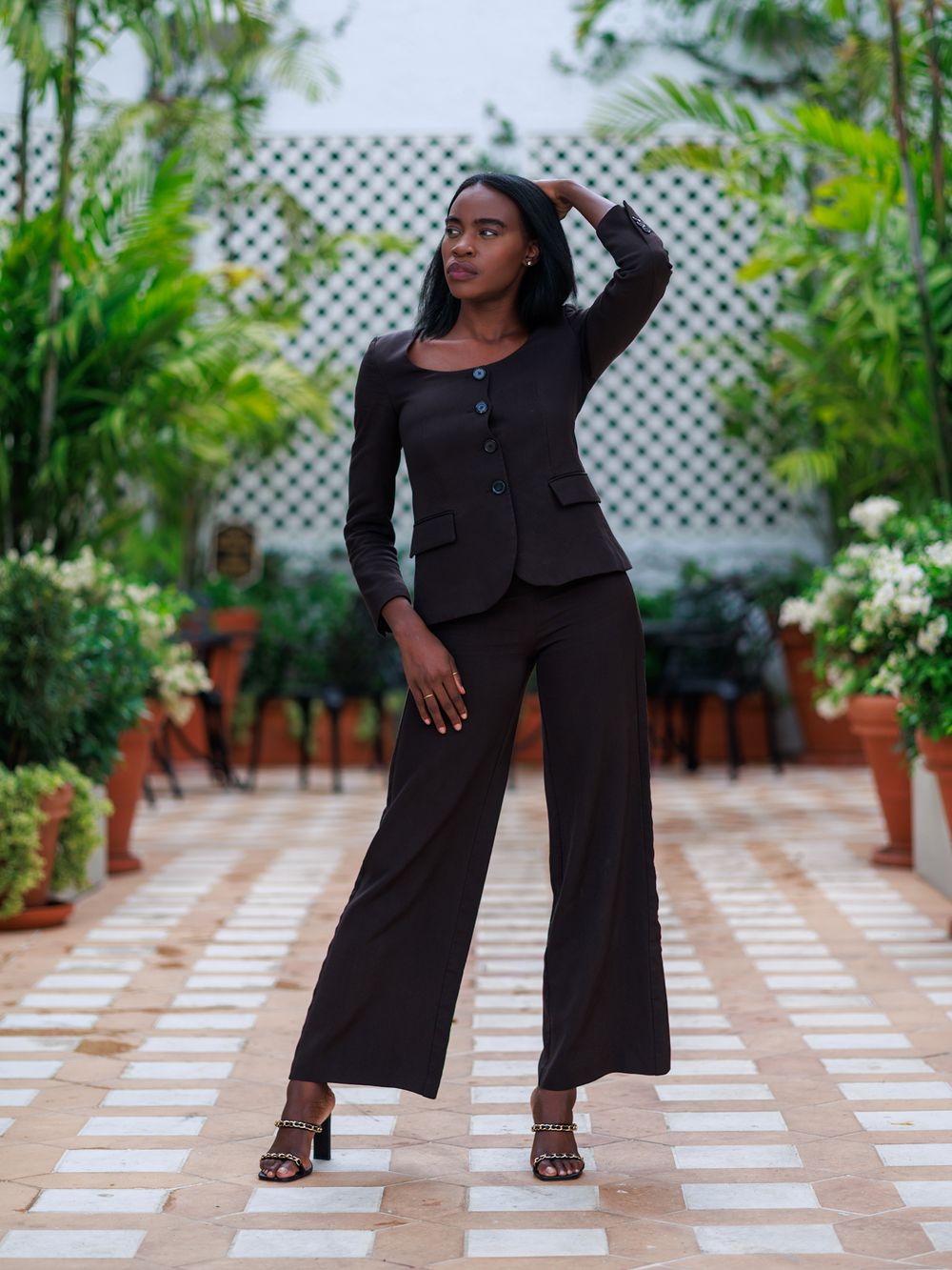 Woman in black blazer and wide-leg pants posing in a garden walkway with lush greenery.