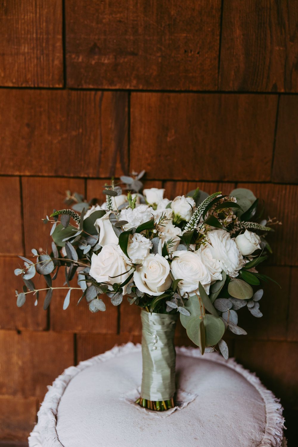 A white rose wedding bouquet with eucalyptus leaves rests on a lace-textured white surface against a rustic wood wall.