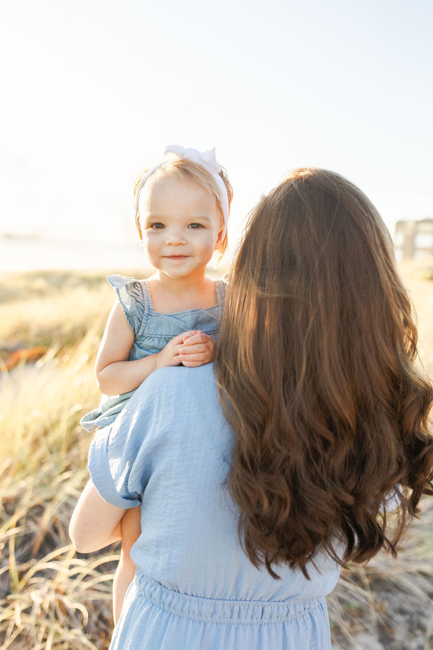 A baby in light blue dress smiles during a golden hour photoshoot in a grassy outdoor setting.Film by Chelsee Rawe Photography