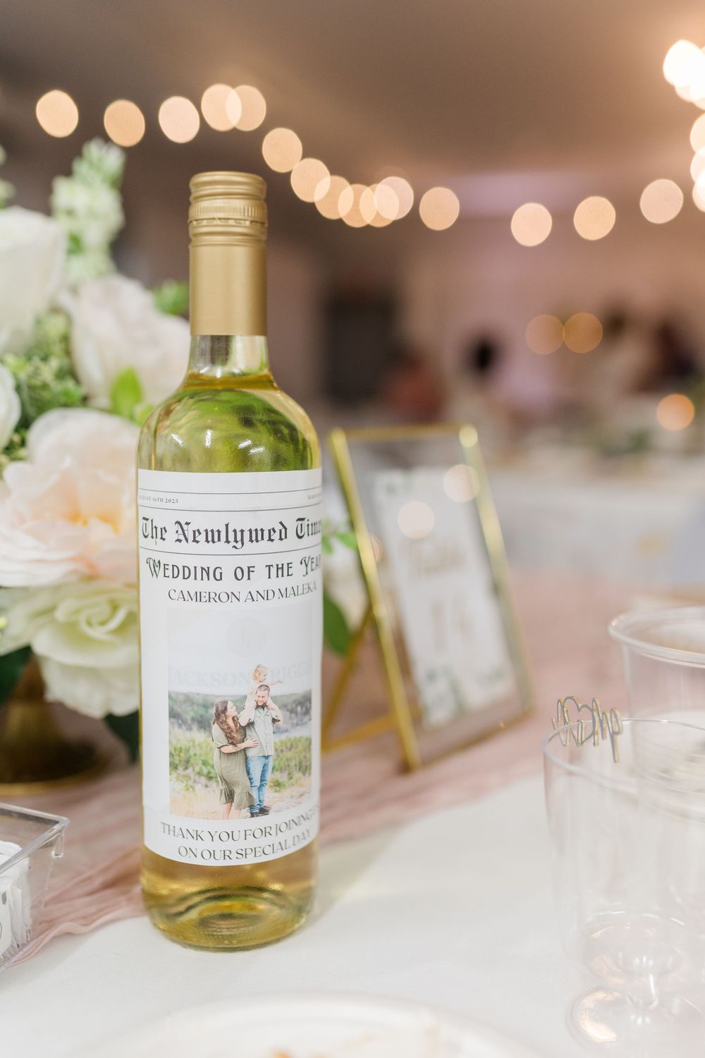 Custom wine bottle labels in gold and cream colors create an elegant table setting display at a wedding reception.