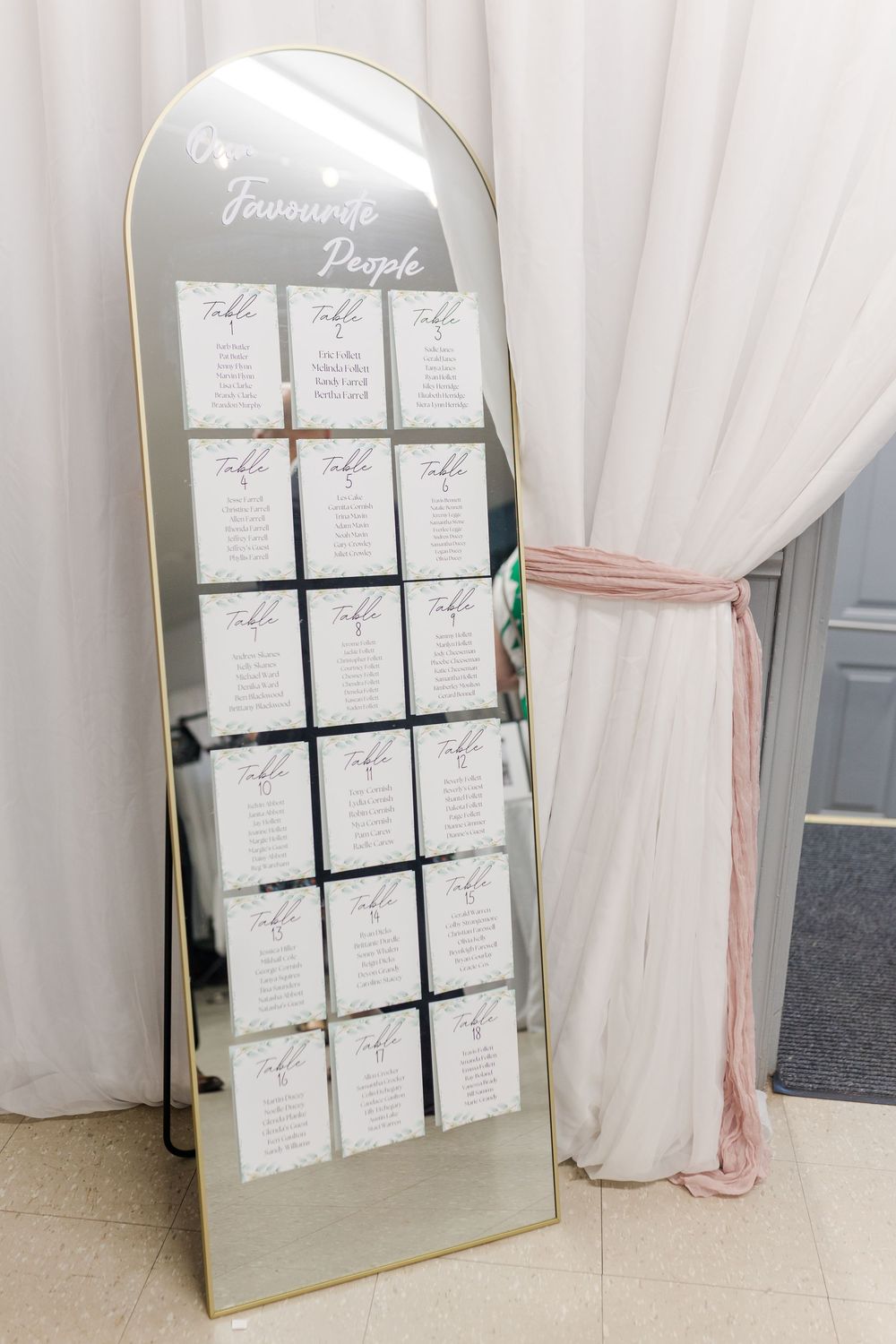 Elegant arched mirror displays wedding seating chart with white cards arranged in rows against a light draped fabric backdrop.