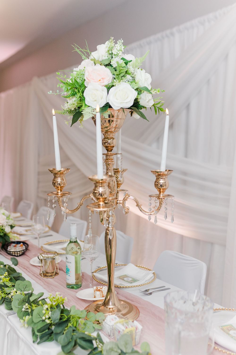 Gold candelabras with white candles and floral arrangements adorn elegant wedding reception tables with a soft pink glow.