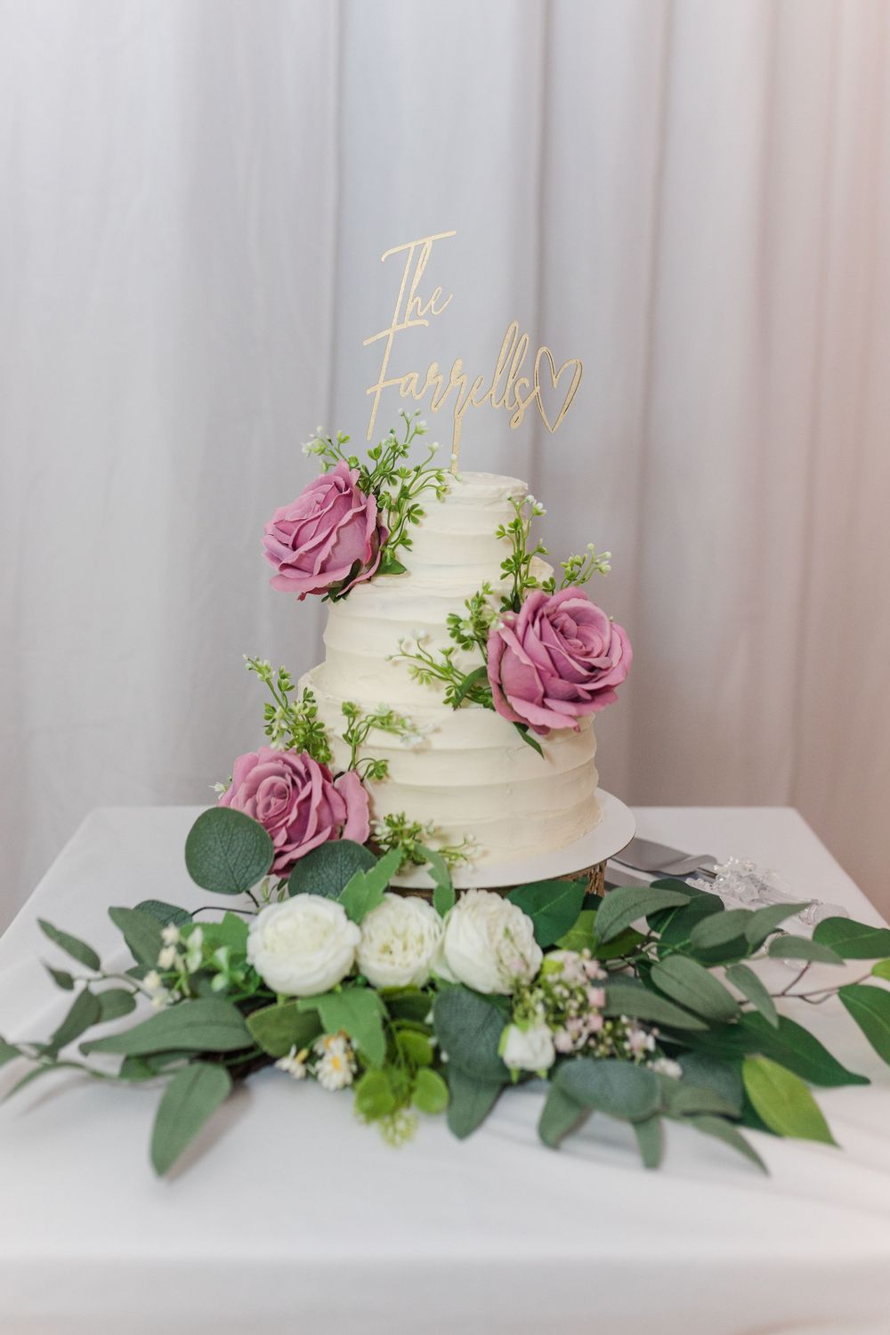 White wedding cake adorned with dusty rose flowers and fresh greenery against a soft backdrop.