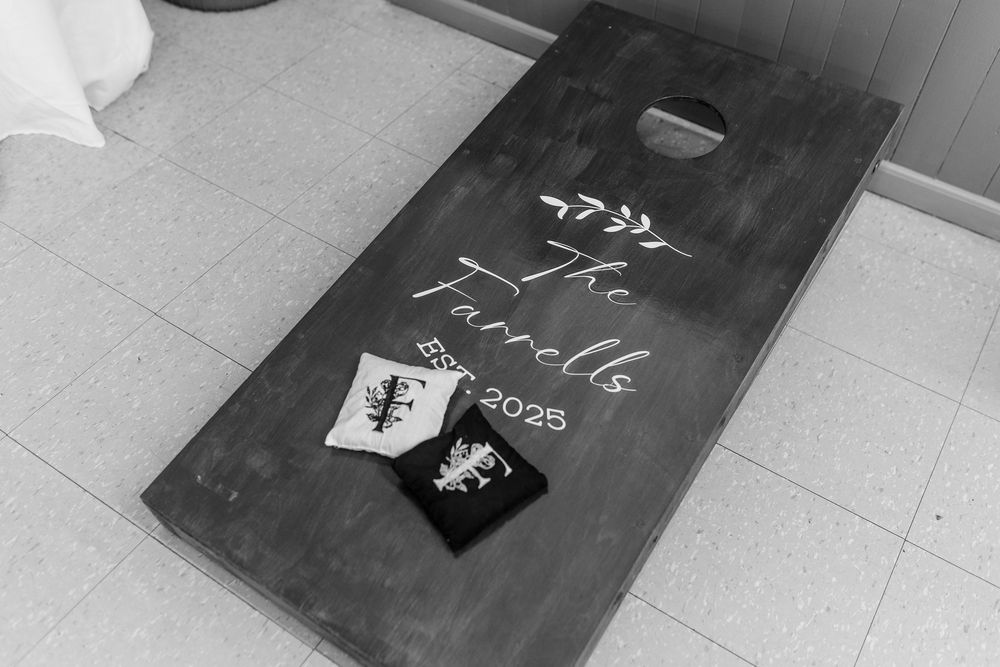 Black and white photograph of a cornhole board with 2023 writing and two bean bags on a tiled floor.