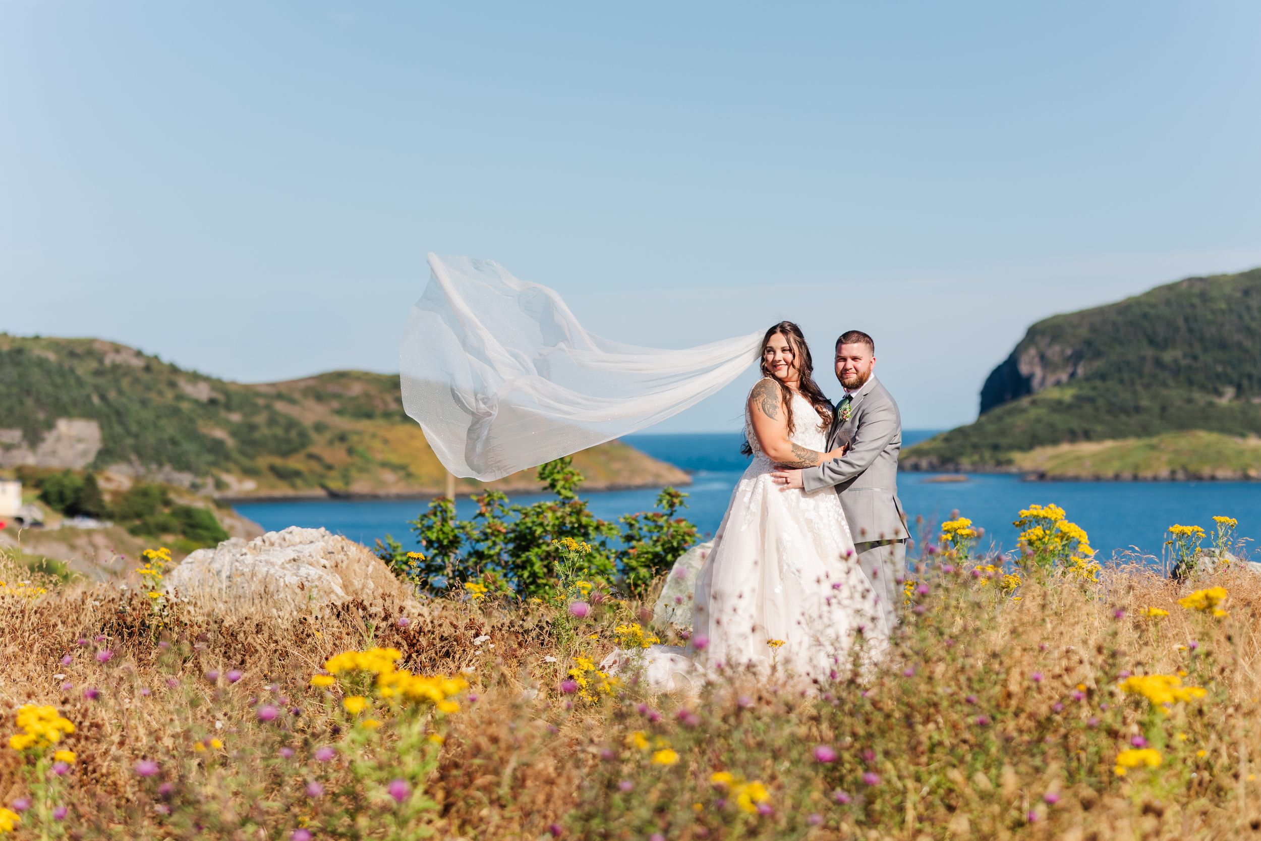 Bride and groom embrace in a beautiful coastal landscape with yellow wildflowers and ocean views.